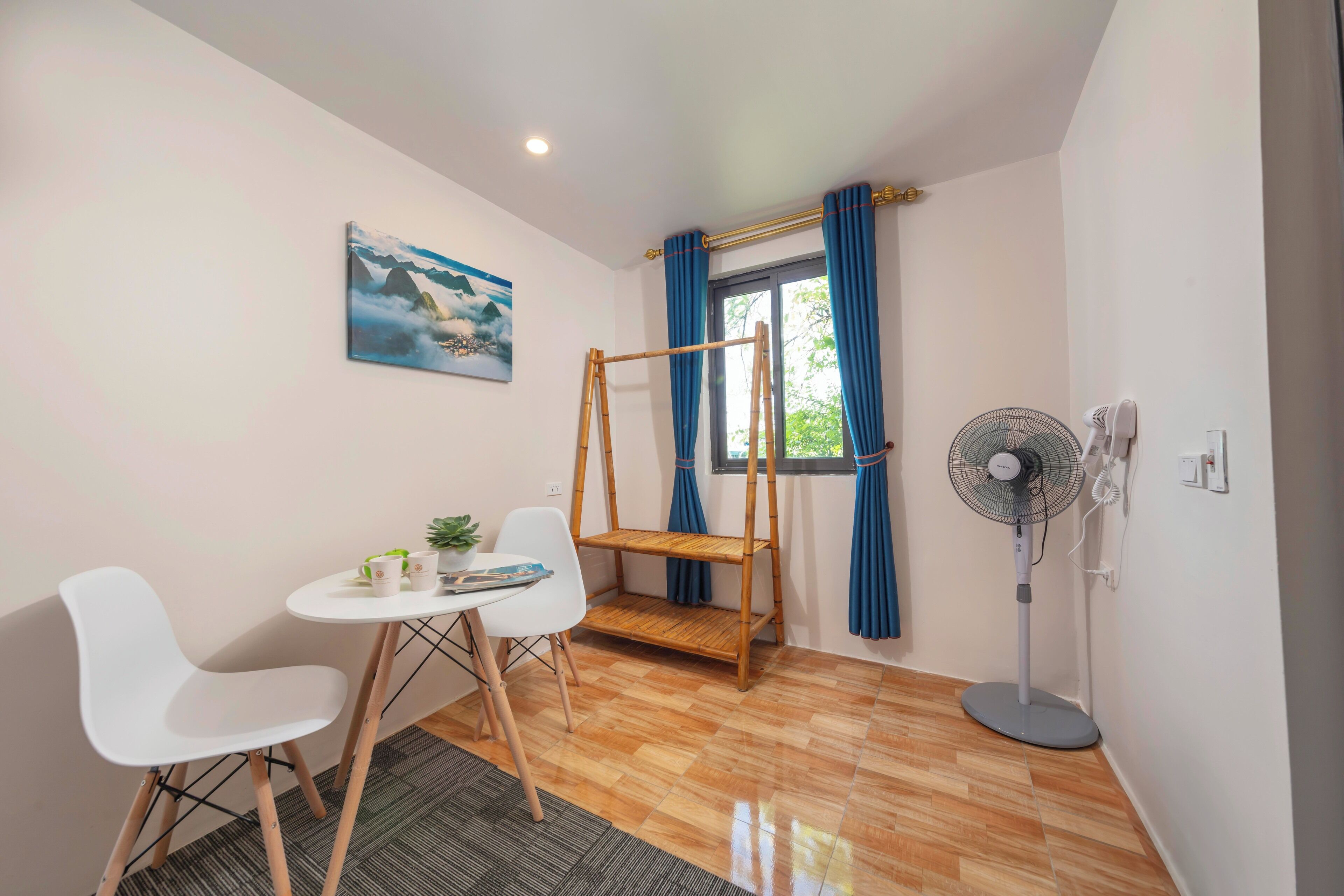 A small room with a wooden floor, a white table with two chairs, a small shelf, a fan, and a window with blue curtains.