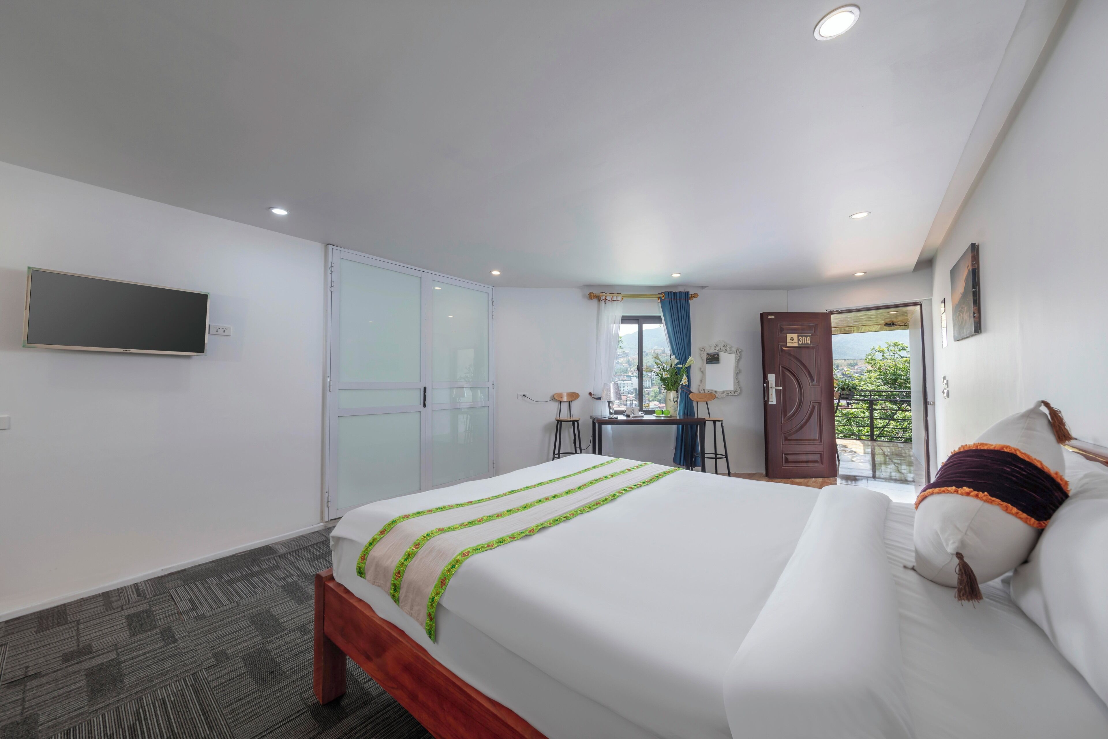 Habitación de hotel con cama, escritorio, silla, televisor, armario y puerta que da a un balcón.