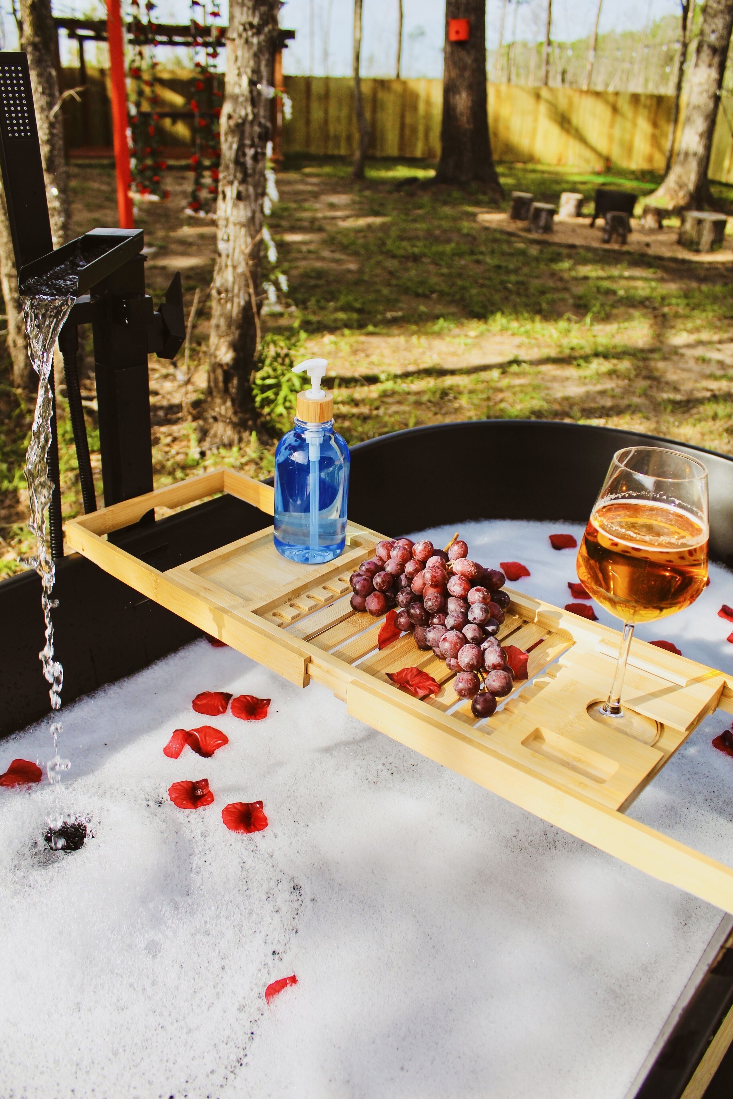 A wooden tray with a bottle of water, a glass of wine, and grapes on a table with rose petals.