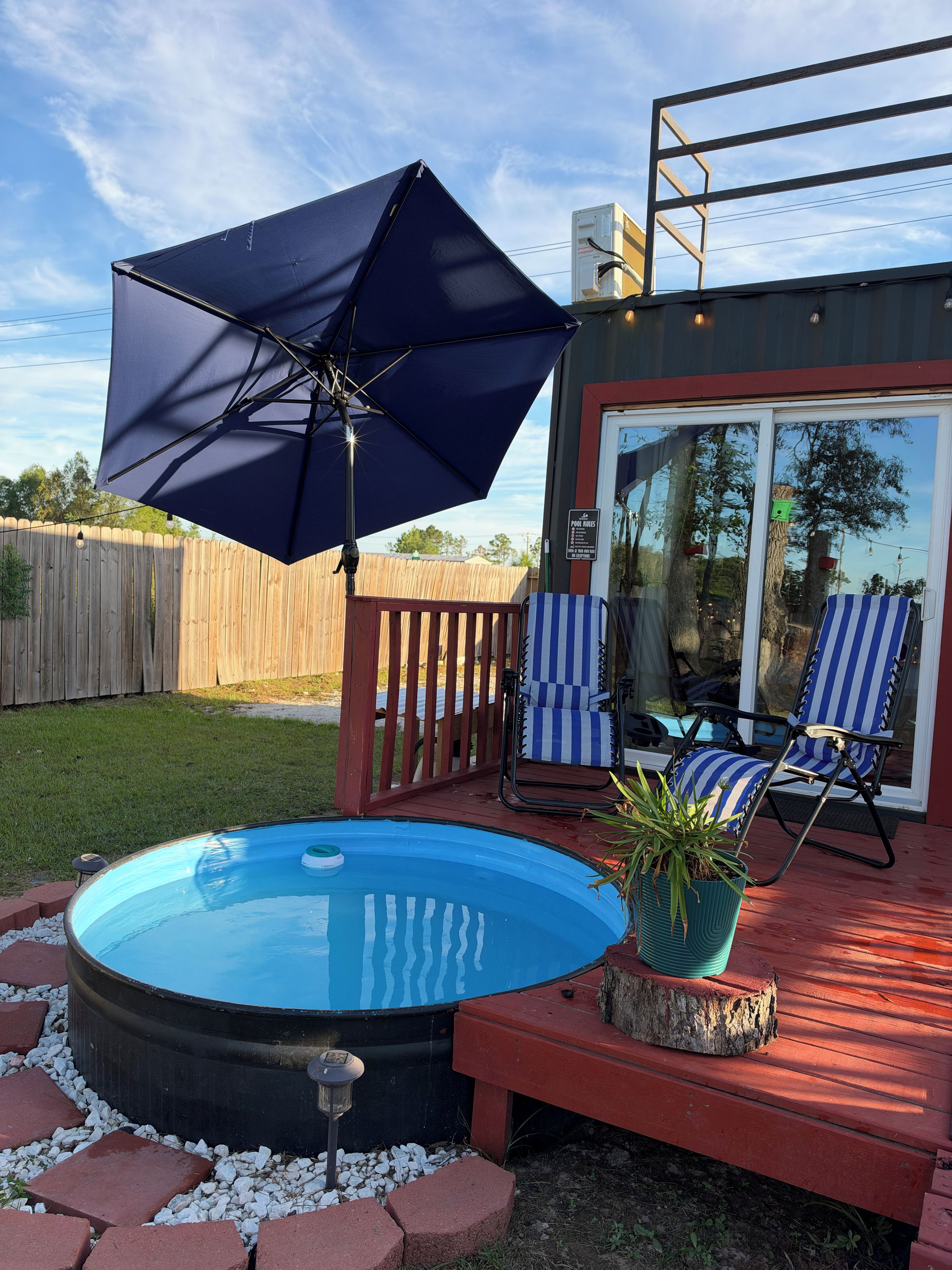 Una piscina al aire libre con una sombrilla azul, una terraza de madera y dos sillones reclinables a rayas.