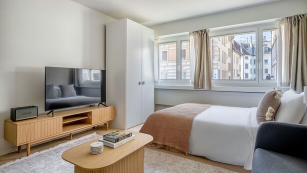 Una habitación de hotel moderna con una cama, un televisor, una mesita de madera y una ventana con cortinas.