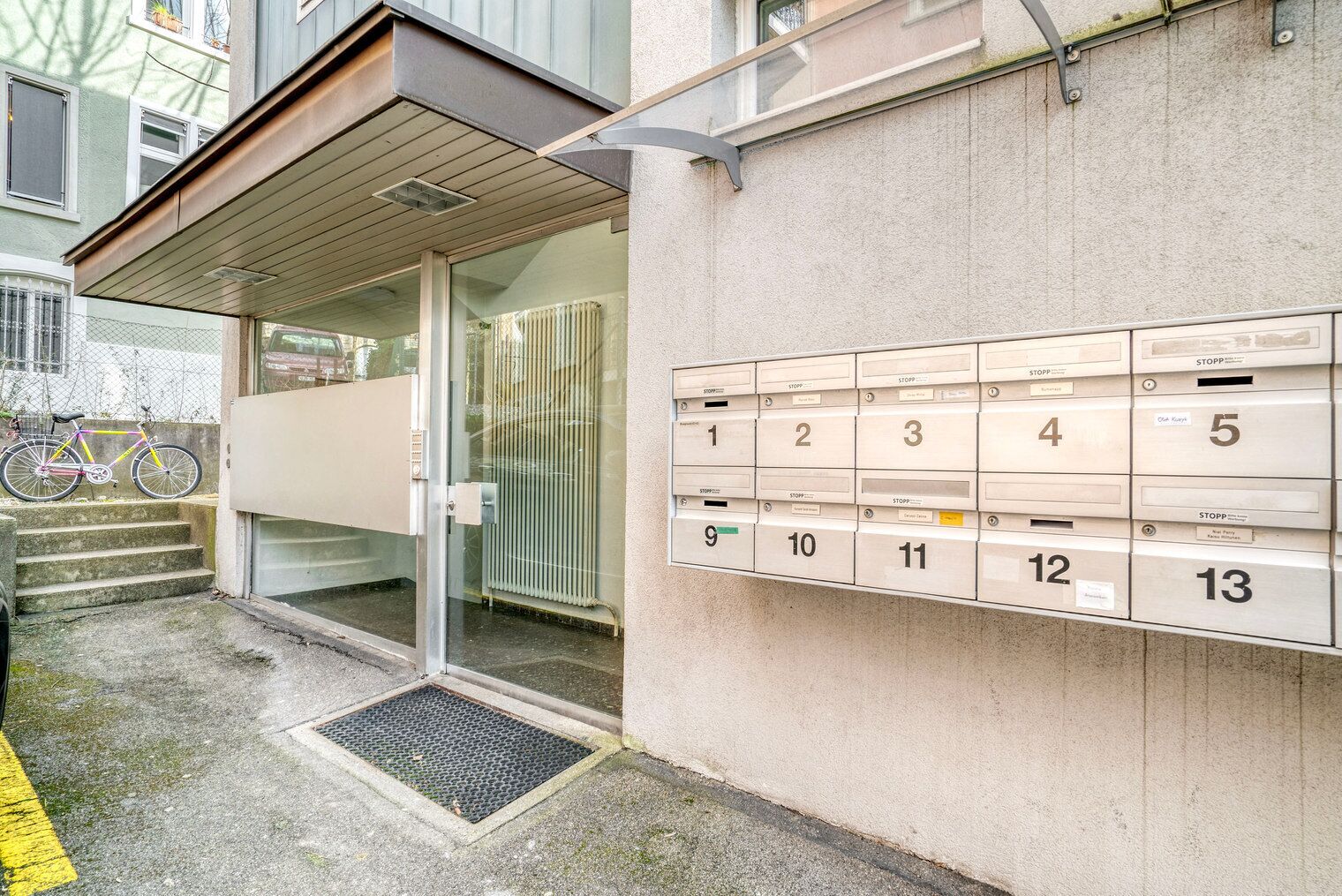 Rakennuksen sisäänkäynti, jossa on lasiovi, ulkona pyörä ja seinällä rivi numeroituja postilaatikkoja.