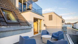 A modern building with a glass balcony, a door leading to an interior room, and a seating area with blue chairs and a round table.