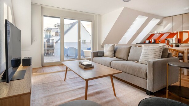 A modern living room with a sofa, coffee table, and a TV. There are large windows and a view of the outside.