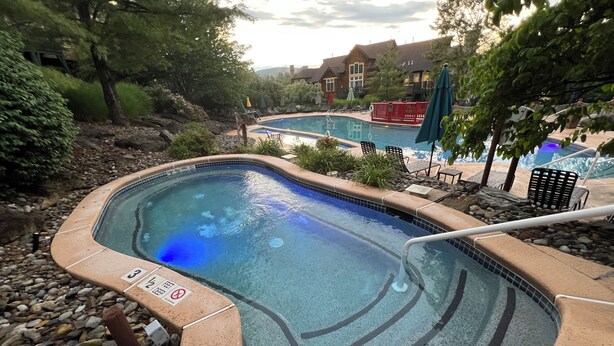 Una piscina con borde de azulejos, rodeada de rocas y plantas, con sillones de descanso y sombrillas cerca.
