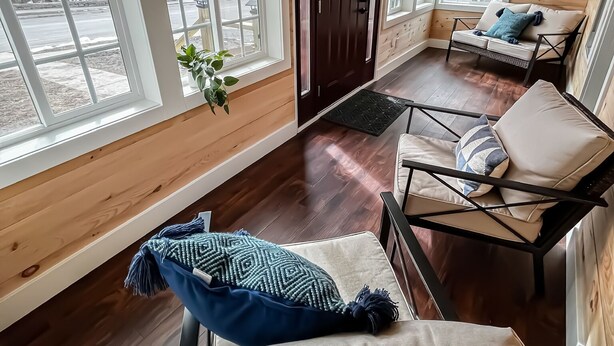 Interior acogedor de una cabaña con paredes y detalles de madera, ventanas amplias, una puerta de madera oscura y una zona de estar con cojines.