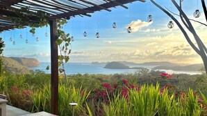 An outdoor dining area with a view of the ocean and hills, covered by a metal roof, and equipped with hanging light fixtures. - Lobster Bay Resort  (Mertak)