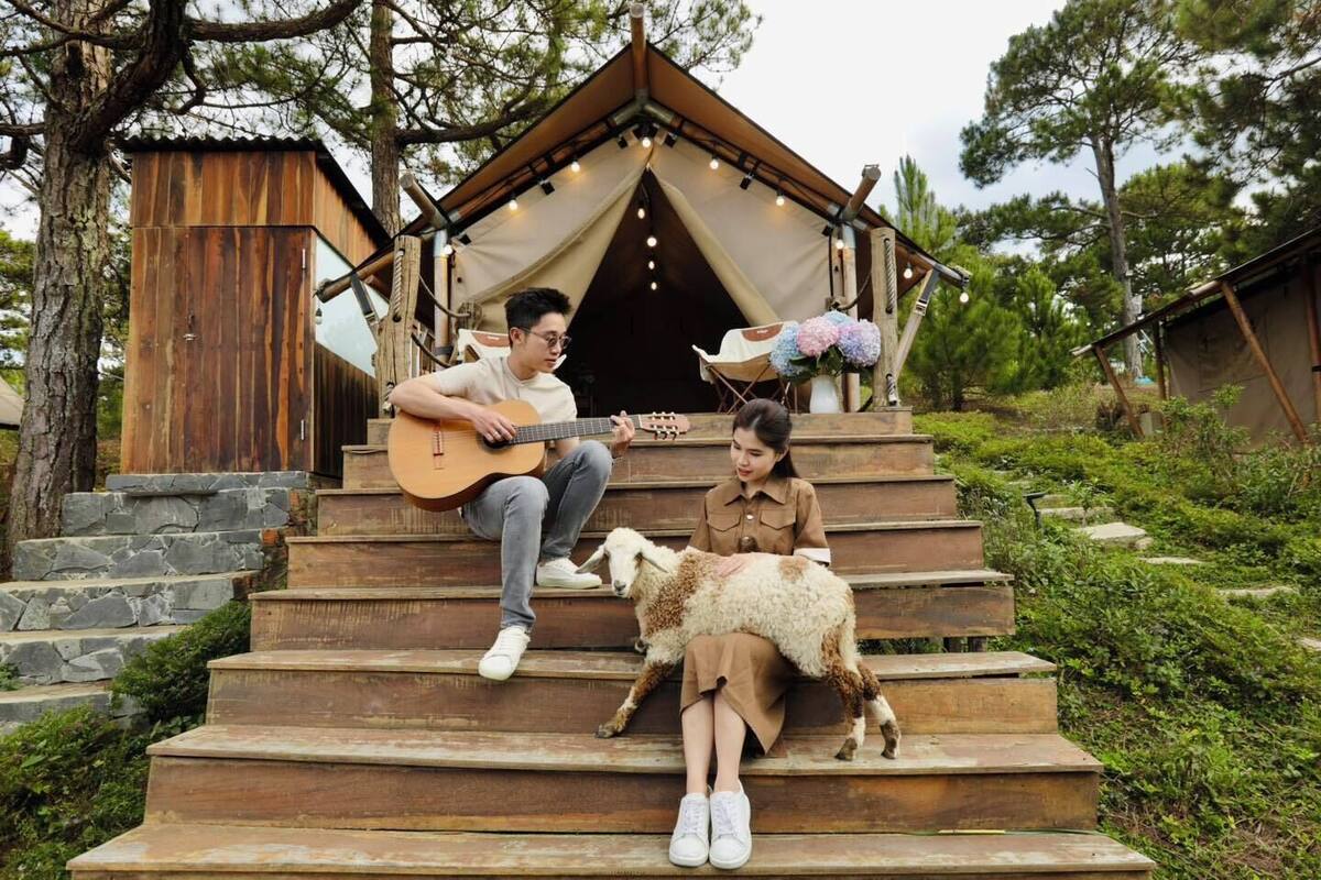 Un escalier en bois rustique mène à une structure en forme de tente où une personne joue de la guitare et une autre est accompagnée d’un mouton.