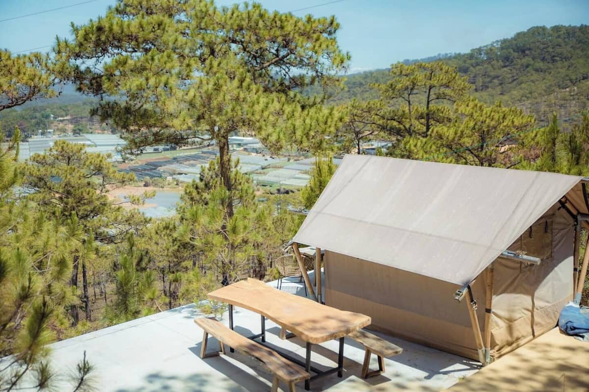 Une tente avec une table et un banc, entourée de pins et offrant une vue sur des champs en terrasses.
