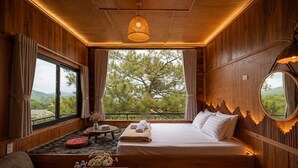 Una habitación acogedora con una cama, un espejo redondo, una ventana con cortinas, una pequeña mesa con un jarrón de flores y vistas a árboles desde el exterior.