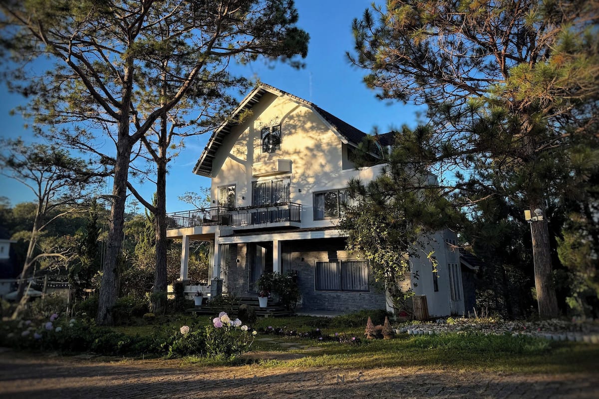 Une maison à deux étages avec un balcon, entourée d’arbres et d’un jardin.