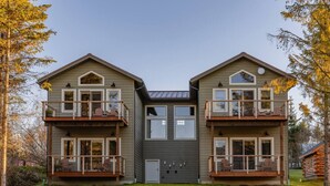 A two-story building with balconies, a central door, and a log cabin in the background. - Lakeshore Lodging Luxury Suites (Homer)