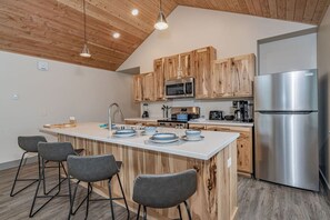 A modern kitchen with wooden cabinets, a stainless steel refrigerator, and a central island with bar stools. - Lakeshore Lodging Luxury Suites (Homer)