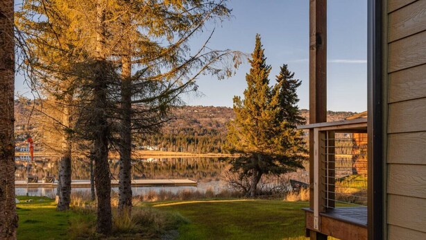 A view from a window overlooking a lake, trees, and a distant building. - Lakeshore Lodging Luxury Suites (Homer)