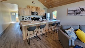 A modern kitchen with wooden cabinets, a central island with bar stools, and a dining area with a gray sofa and patterned cushions. - Lakeshore Lodging Luxury Suites (Homer)