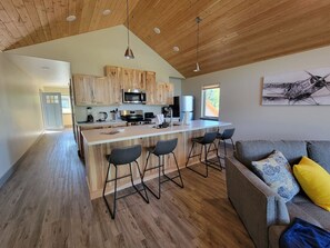 A modern kitchen with wooden cabinets, a central island with bar stools, and a dining area with a gray sofa and patterned cushions. - Lakeshore Lodging Luxury Suites (Homer)