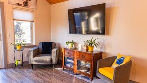 A living room with a sofa, armchair, wooden side table, and a TV mounted on the wall. - Lakeshore Lodging Luxury Suites (Homer)
