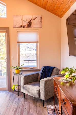 A cozy room with a grey armchair, a wooden cabinet, and a window with blinds. - Lakeshore Lodging Luxury Suites (Homer)