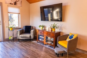 Een woonkamer met een bank, fauteuil, houten bijzettafeltje en een aan de muur bevestigde televisie.