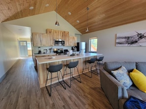 A modern kitchen with wooden cabinets, a central island with bar stools, and a dining area with a grey sofa and patterned cushions.