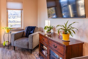 Uma sala de estar com uma poltrona cinza, um aparador de madeira com gavetas, uma mesa pequena com uma planta em vaso e uma janela grande com vista para árvores.