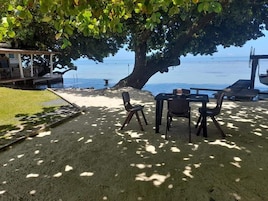 Un área de descanso frente a la playa con una mesa y sillas bajo un árbol grande, con vistas al mar.