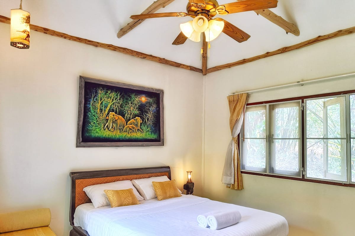 A bedroom with a bed, a ceiling fan, a window with curtains, and a painting on the wall.