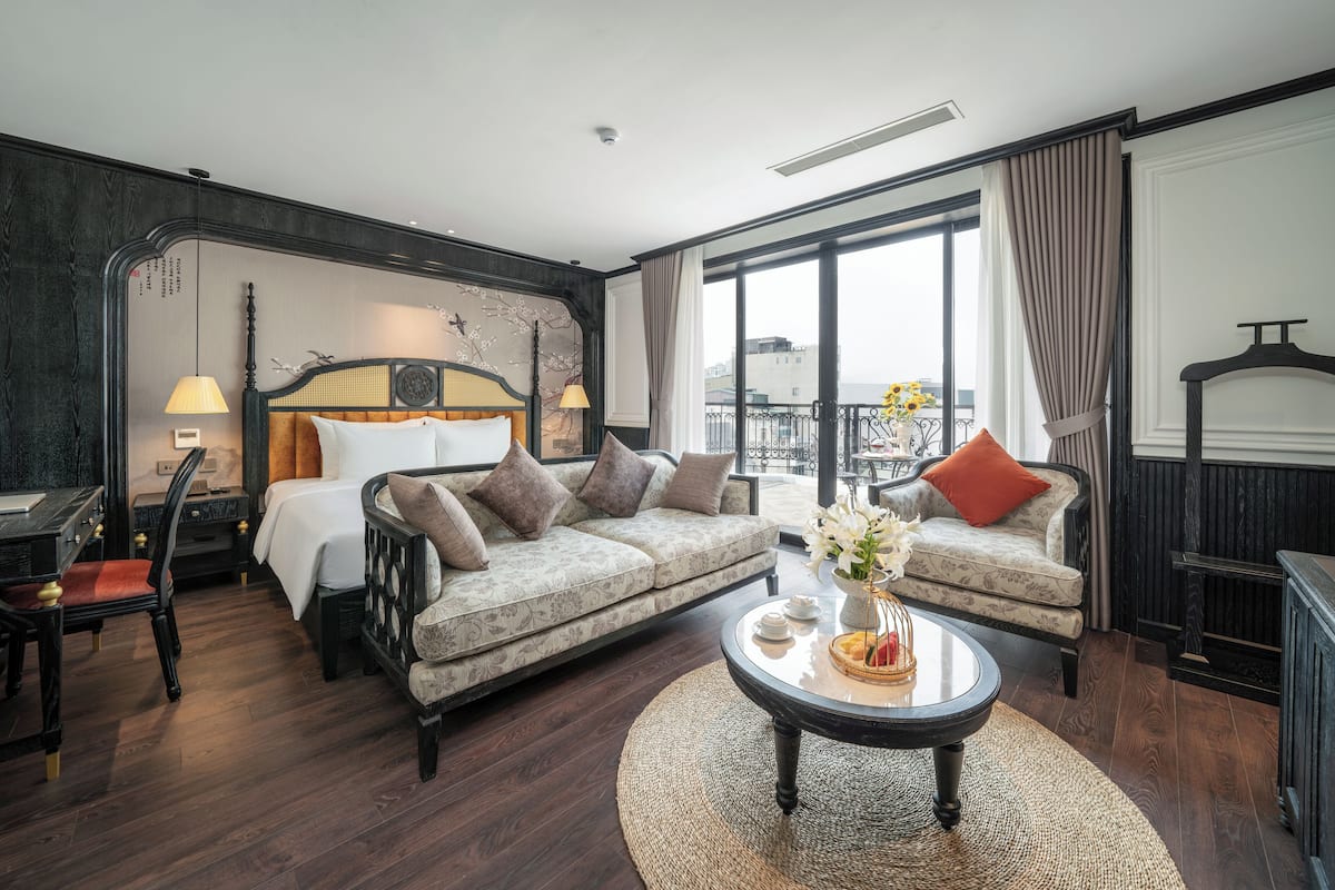 Moderni hotellihuone, jossa on suuri sänky, sohva, pyöreä sohvapöytä ja suuret ikkunat, joista avautuu näkymä kaupunkiin.