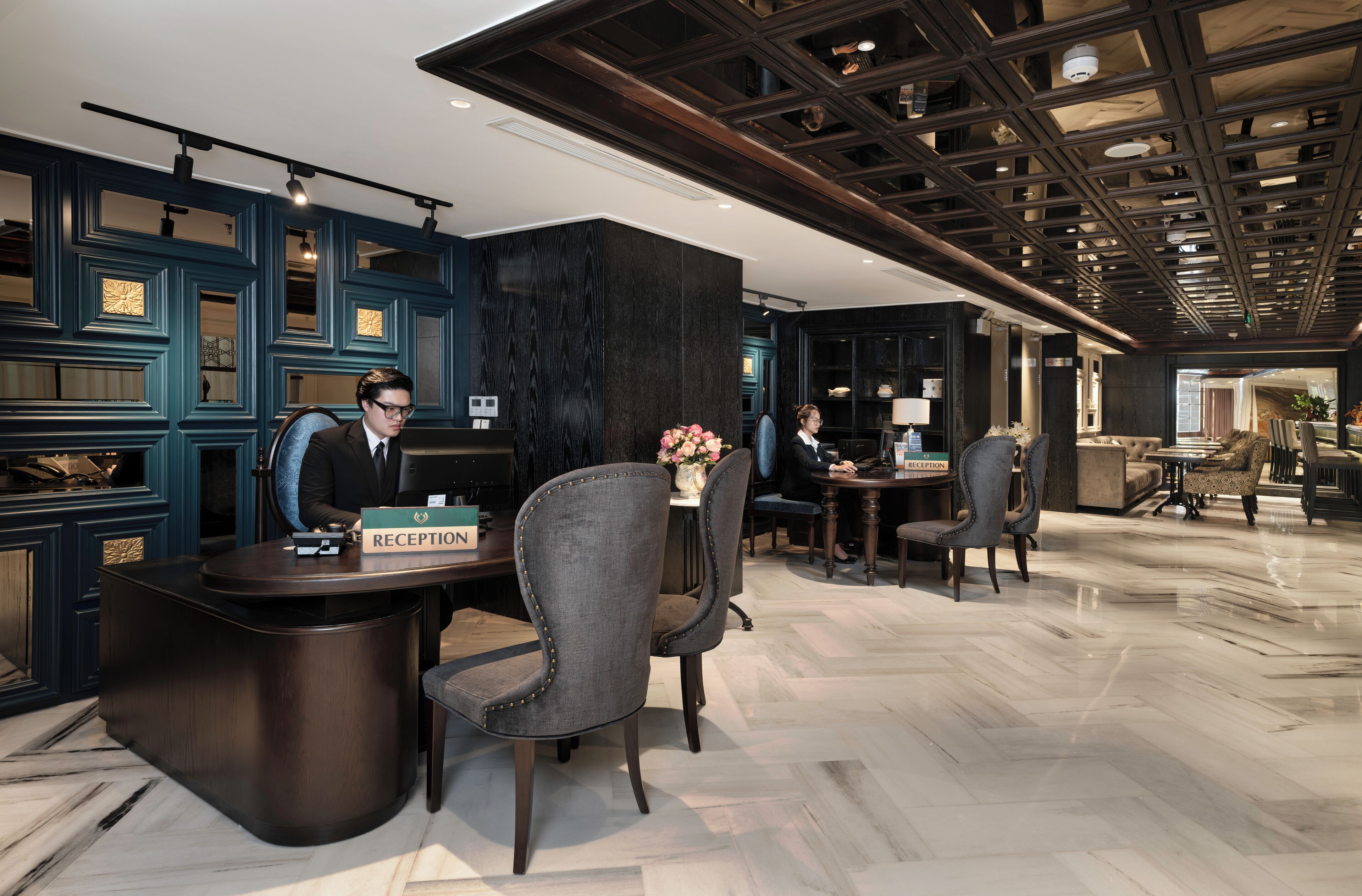 A reception area with a reception desk, two chairs, a vase with flowers, and a dark wooden wall with decorative panels.