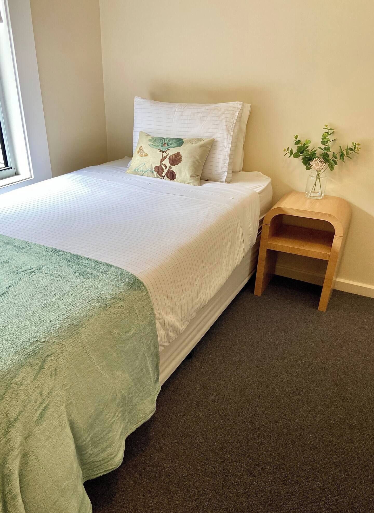 A neatly made bed with a green blanket, a wooden nightstand with a vase of flowers, and a window to the left.