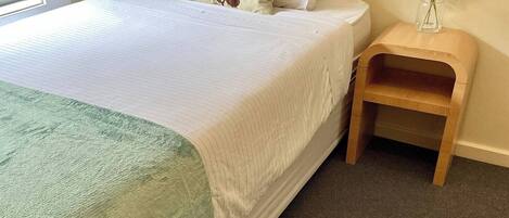 A neatly made bed with a green blanket, a wooden nightstand with a vase of flowers, and a window to the left.