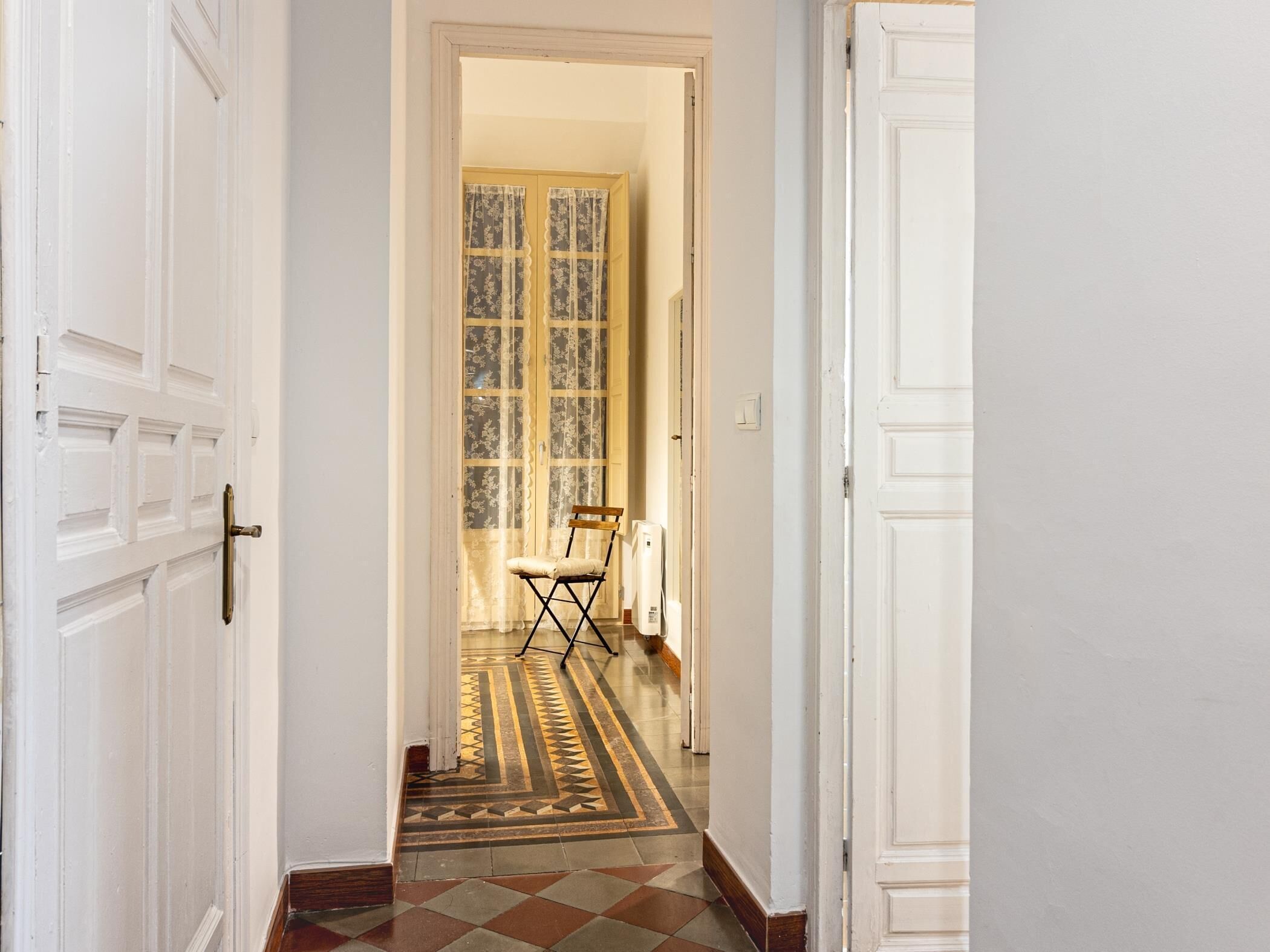 Un couloir avec des portes blanches, un sol à motifs et une chaise au loin.