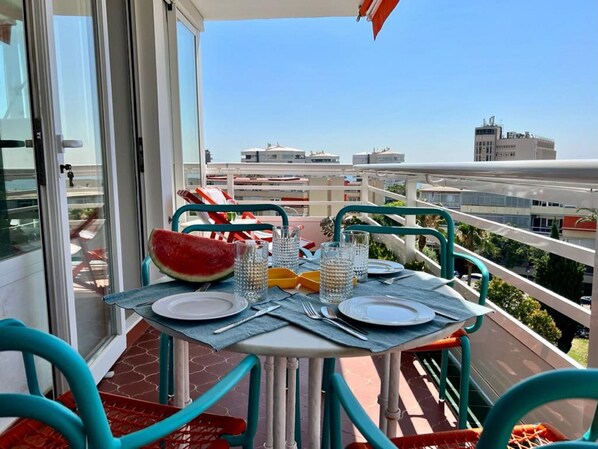 Ein Balkon mit Tisch, Geschirr, Besteck und einer Wassermelone, mit Blick auf eine Stadtlandschaft.