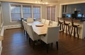 A dining area with a wooden table and white chairs, a kitchen with a bar, and large windows with blinds. - Middletown Bungalow w/Hot Tub and Private Yard - RIBryan Properties (Middletown)