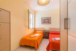 A small hotel room with two single beds, a wooden wardrobe, a bedside table with a lamp, and a painting on the wall.