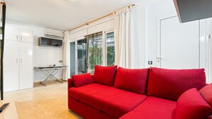 A modern living room with a red sofa, a small table, and a kitchenette area with white cabinets and an air conditioning unit. - WELCS APARTAMENTO 126 PDA cerca del puerto (Platja d´Aro)