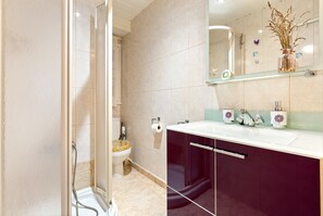 A bathroom with a glass shower enclosure, a toilet, a sink with a mirror above, and a vase with dried flowers on the sink's edge.