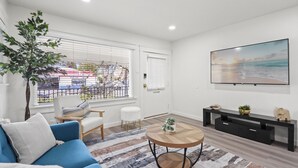 A modern living room with a blue sofa, a round coffee table, a black TV stand, and a large window with blinds. - 1 Mile to the Beach, Games, Remodeled 3BD Home, Fire Pit, Patio (Long Beach)
