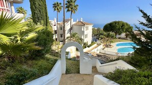 A pathway leading to a pool area with a clear view of the ocean, surrounded by palm trees and a building with a tiled roof. - 17 - sea view Apt. in Calahonda (Mijas Costa)