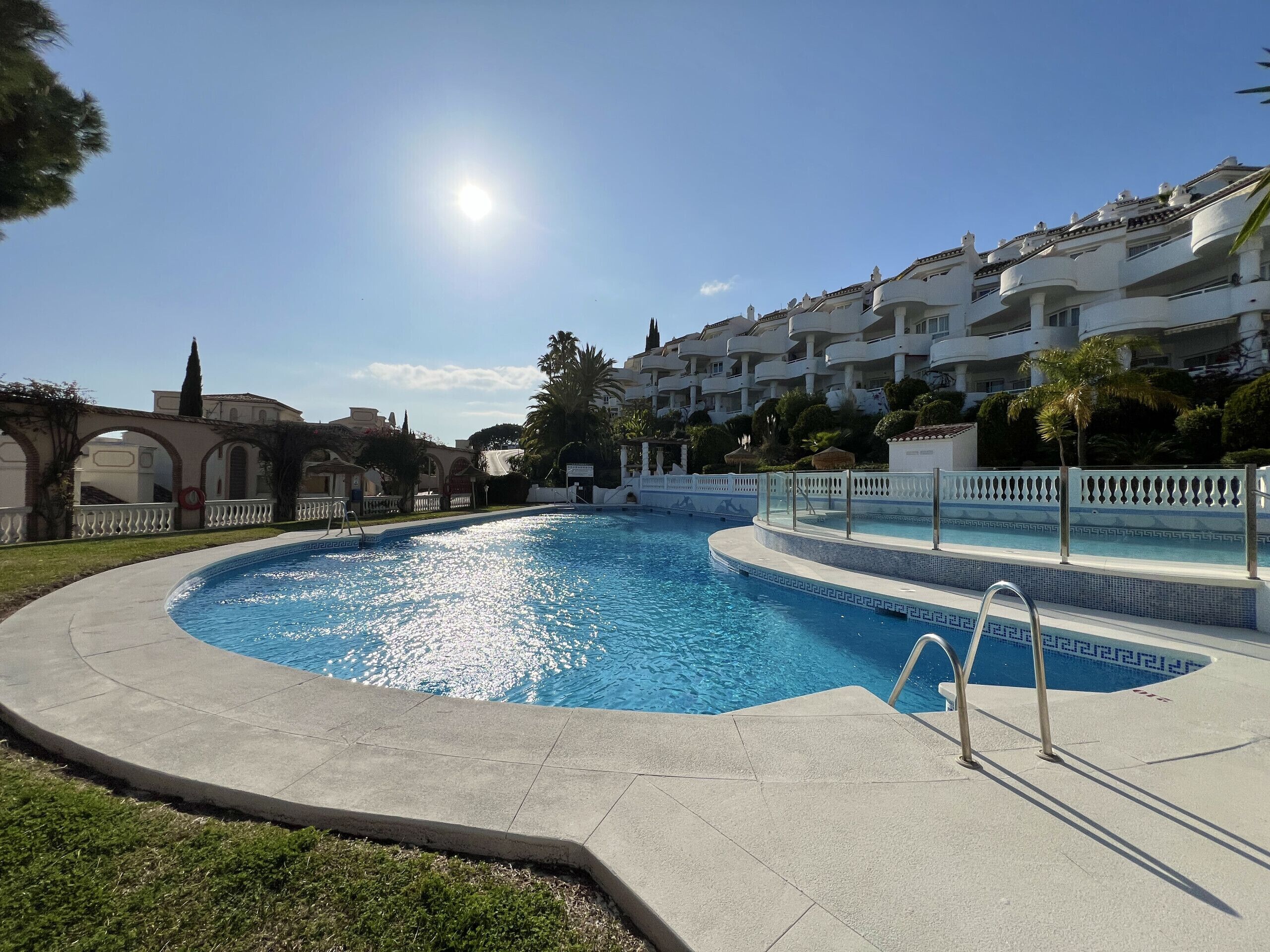 A swimming pool with a clear blue water, surrounded by a white fence and a paved walkway.
