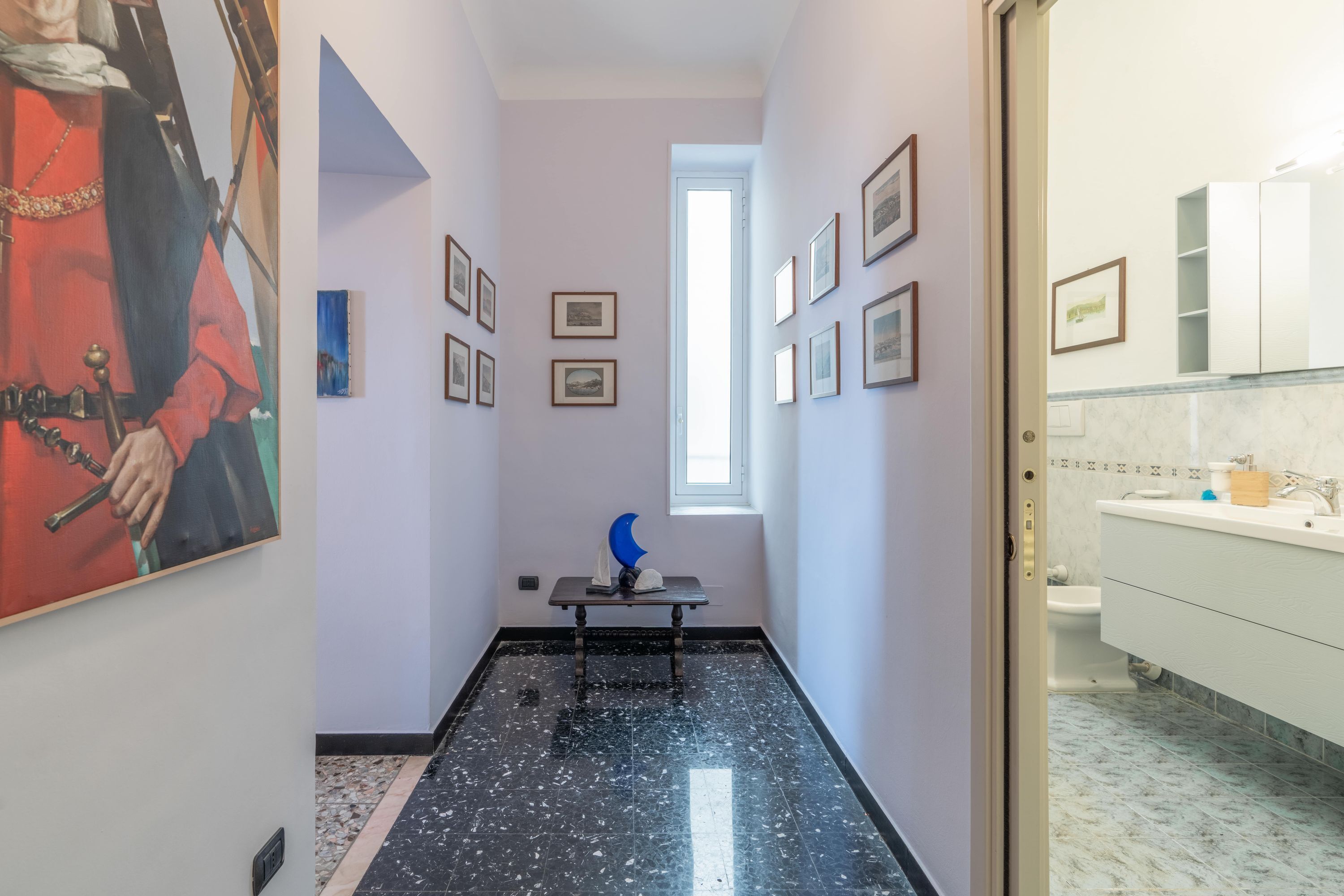 Een gang met een schilderij, ingelijste foto's, een tafeltje met een beeldhouwwerk en een badkamer met een wastafel en spiegel.