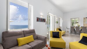 Een moderne woonkamer met een grijze bank en geel stoelen, een eethoek met een tafel en stoelen, en een groot raam met uitzicht op de zee.