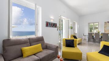 A modern living room with a grey sofa and yellow armchairs, a dining area with a table and chairs, and a large window offering a view of the sea.