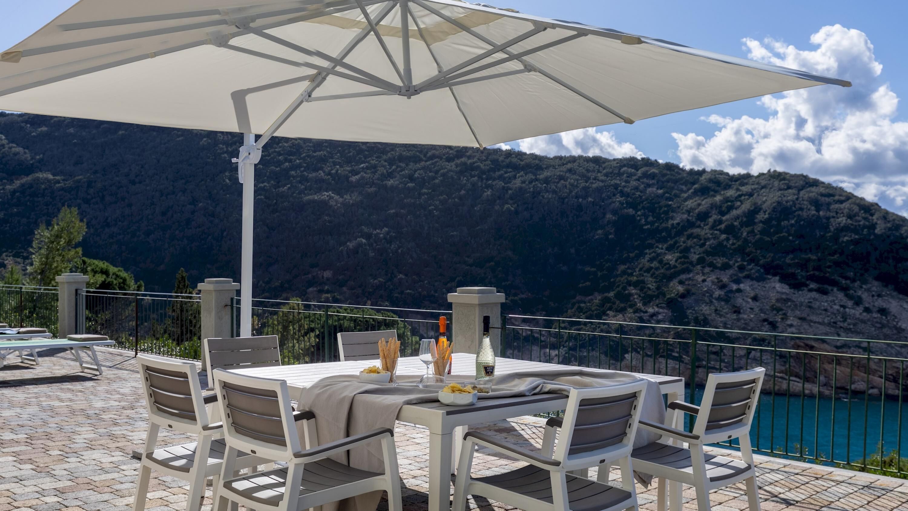 Mesa posta ao ar livre com vista para montanhas, um guarda-sol branco e uma garrafa de vinho.