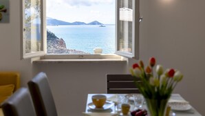 Een eetgedeelte met een tafel gedekt voor twee personen, een raam met uitzicht op zee en een veraf gelegen eiland, en een vaas tulpen op de tafel.