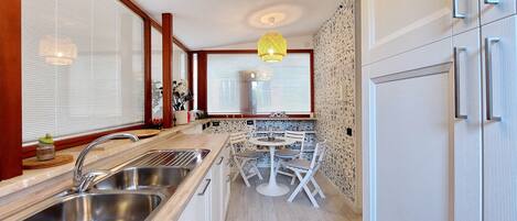 A modern kitchen with a double sink, white cabinetry, a round dining table with chairs, and a window with blinds.
