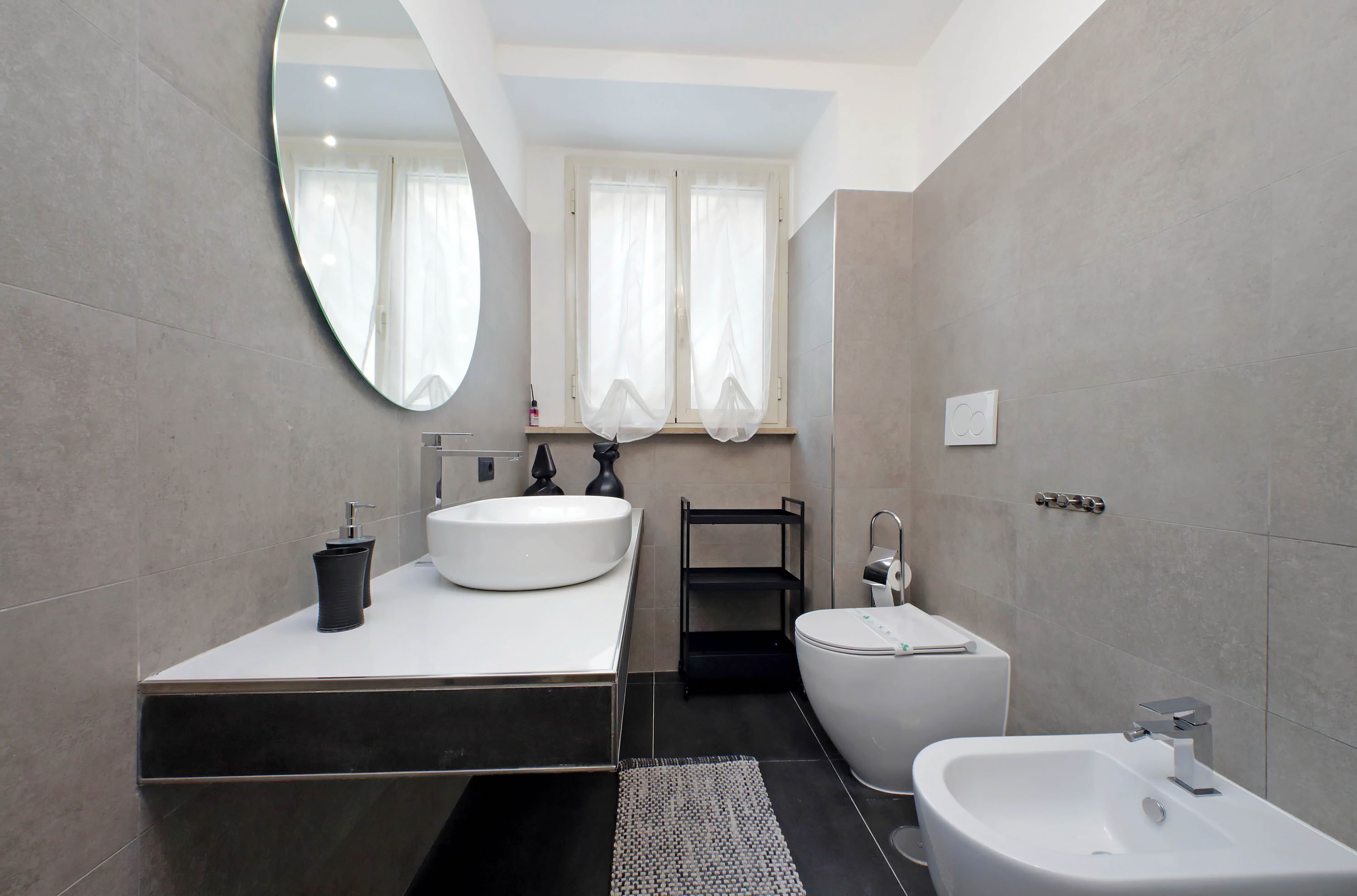 A modern bathroom with a large oval mirror, a white sink, a bidet, and a toilet.