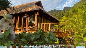 A wooden dining area with a large table and chairs, a potted plant, and a view of a thatched-roof building and lush greenery. - Mơ Village (Da Bac)