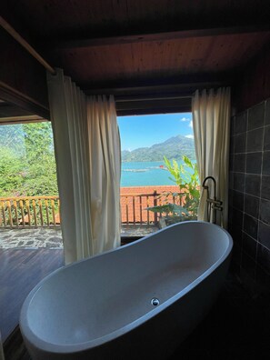 Ein Badezimmer mit freistehender Badewanne, Blick auf ein Gewässer und einer Holzterrasse.