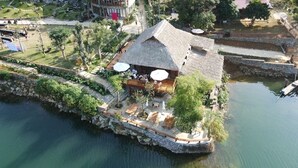 A thatched-roof building with outdoor seating and a view of a calm body of water. - Mơ Village (Da Bac)